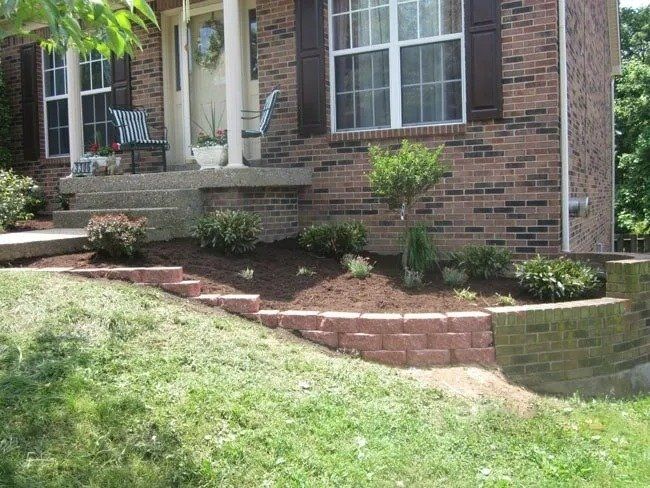 A brick house with a raised flower bed. Brown brick wall with steps, mulch, and green plants.