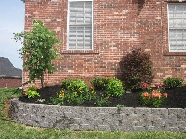 A brick house with a retaining wall supporting a landscaped flower bed of bushes and flowers.
