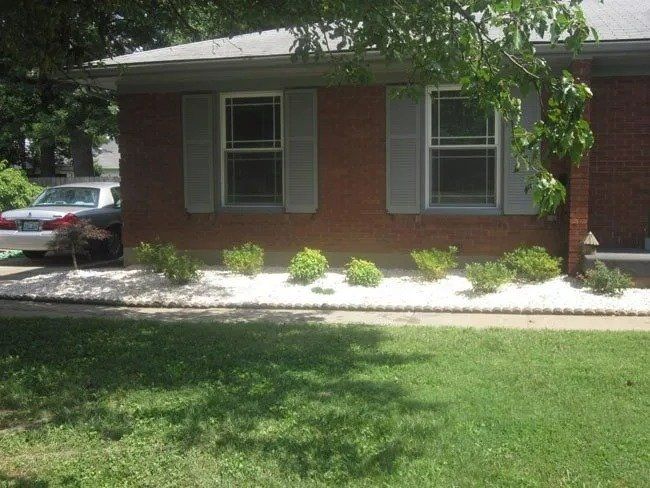 A brick house with gray shutters and a white gravel flower bed. A car is parked beside the house.