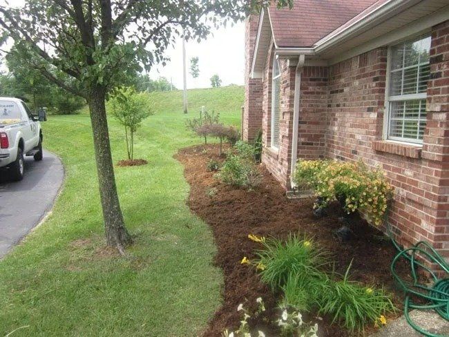A brick house with a mulched flower bed, grass, and a tree. A white truck is parked on the left.