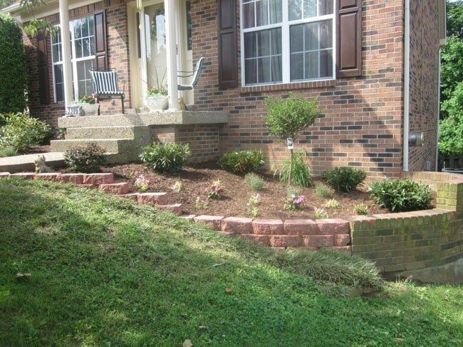 A brick home with a small garden bed, steps, and retaining wall; a grassy yard slopes down from the house.