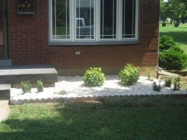 Brick house with a window above a white-rock-filled garden bed, edged with a decorative border, with shrubs and green grass.