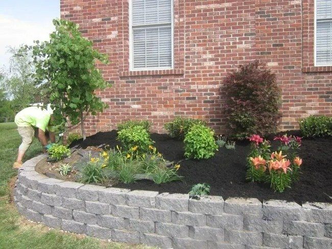 A person planting flowers in a landscaped bed with a low retaining wall in front of a brick house.