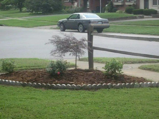 A landscaped front yard with a small tree and bushes in a mulch bed, bordered by stone. A car sits on the street in the background.