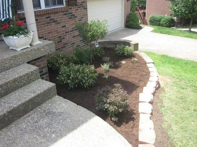 Brick house with a small garden bed bordered by stone edging. Mulch and various green bushes fill the bed next to the steps.