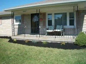 A small, brick home with a porch and decorative ironwork. A bench sits on the porch, and there is a small yard in front.