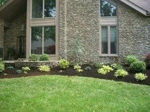 House exterior with stone facade and landscaped bed, featuring various shrubs and fresh mulch, alongside a green lawn.