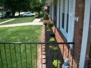 View of a brick house with a garden bed alongside. Black railing in the foreground. Lush green lawn in the background.