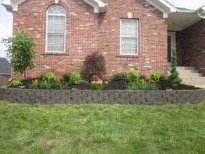 Brick house with a low dark retaining wall filled with plants. A green lawn is in the foreground.