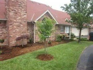 A brick home with a red roof, green lawn, and newly planted trees. Brown mulch borders the lawn.