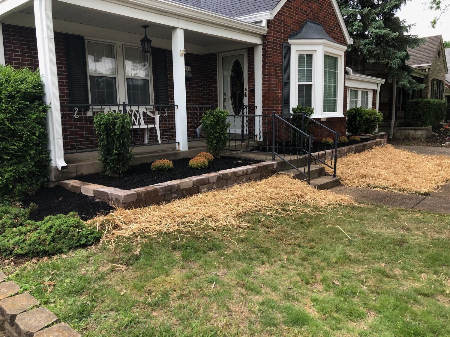 A brick house with a porch and bay window, featuring a landscaped front yard with mulch and low-lying shrubs.