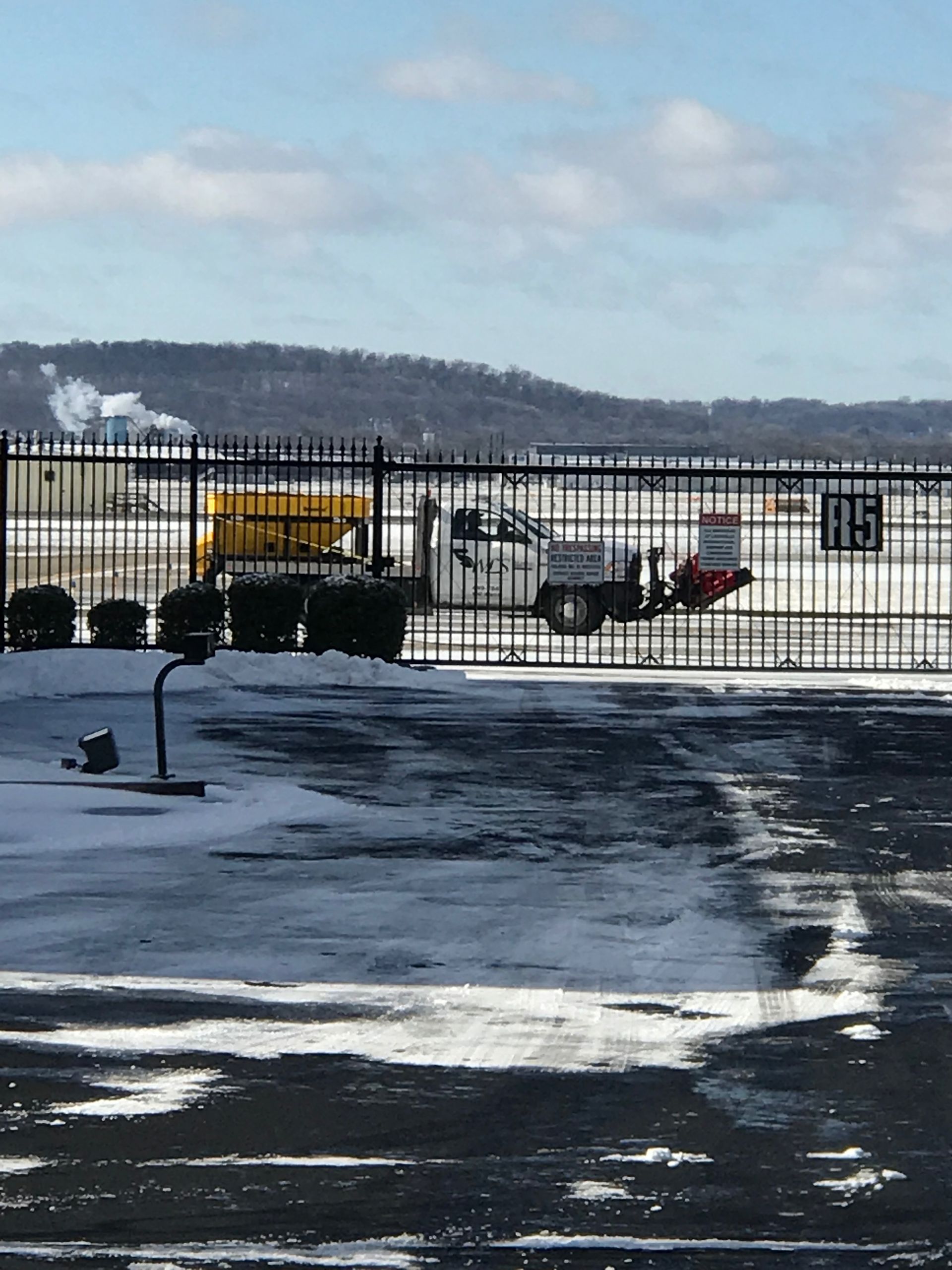 Snowy scene at a gated area. A pickup truck with a snowplow is behind the gate. Background includes a hill and smoke stacks.