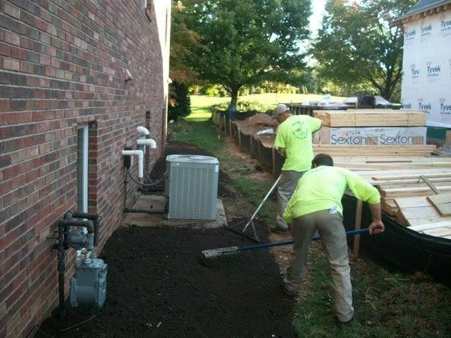 Two workers in green shirts rake dark soil along the side of a brick building, near an air conditioner.
