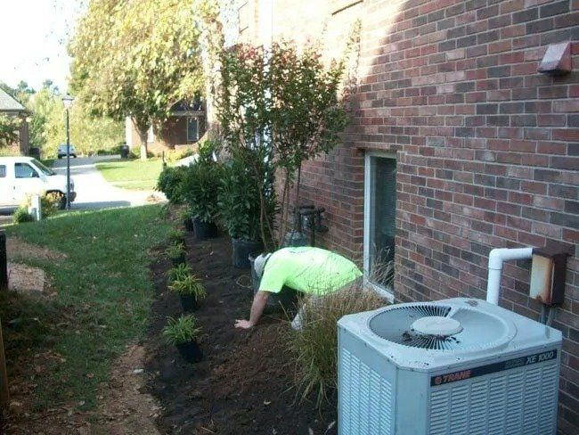 Man planting small plants in a garden bed next to a brick building and an air conditioning unit.