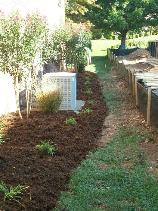 Landscaped backyard with mulched flowerbeds, an air conditioning unit, and a narrow grass path next to a retaining wall.