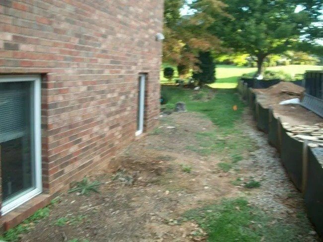 Brick building exterior with windows, dirt, and a retaining wall along a grassy area. A tree is visible in the distance.