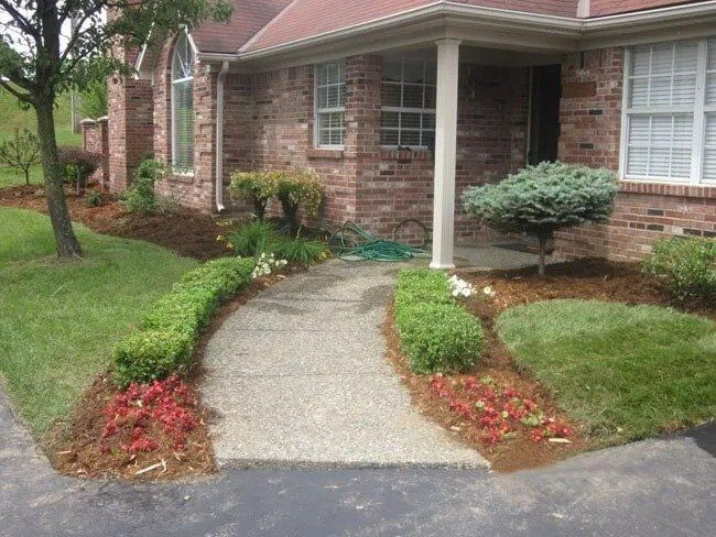 A brick house with a concrete walkway, landscaped with red flowers, green bushes, and brown mulch.