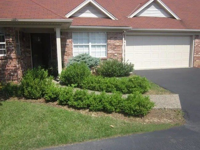Exterior of a brick townhouse with a red roof and garage. Green shrubs line the front, with a walkway and grass in front.
