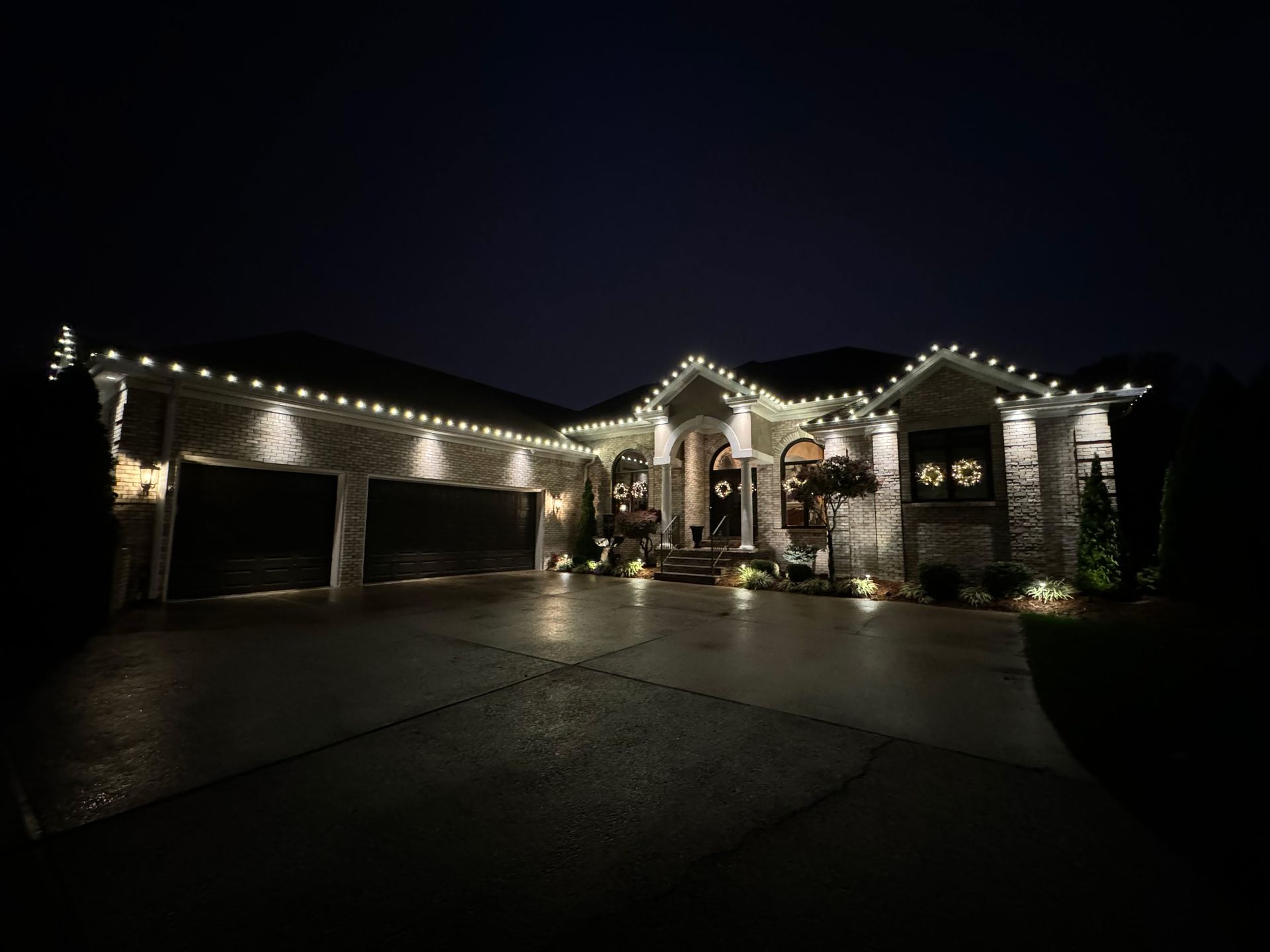 A house at night, decorated with white Christmas lights along the roofline and other areas. Driveway reflects the lights.