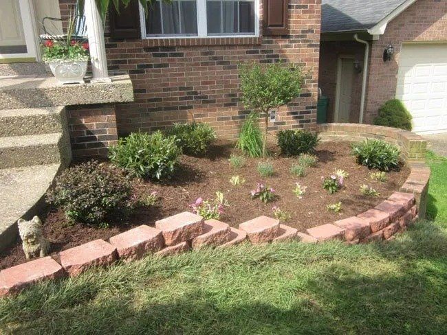Red brick retaining wall encloses a flower bed with various green plants and mulch, in front of a brick house.