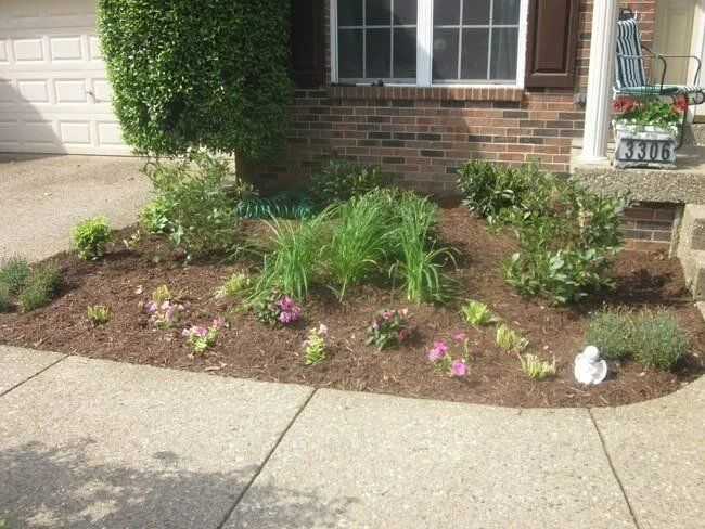 A front yard flower bed with fresh mulch, various plants, and a small white statue against a brick house.