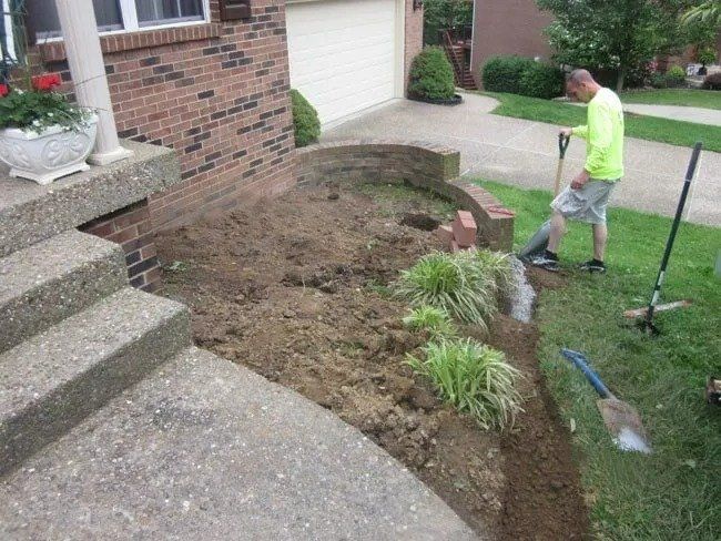 Man digging in a yard next to a brick wall and steps. Dirt is exposed in a garden bed with some plants.