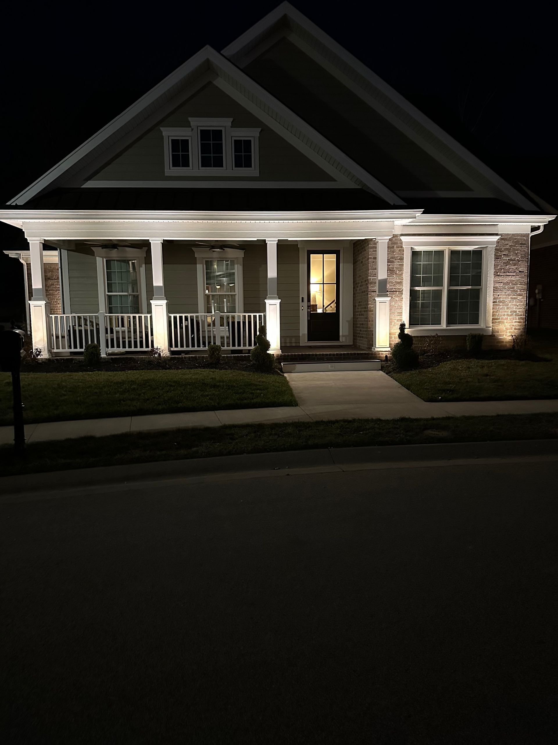A house lit up at night, featuring a porch and sidewalk. Beige siding with white trim, and a dark roof.