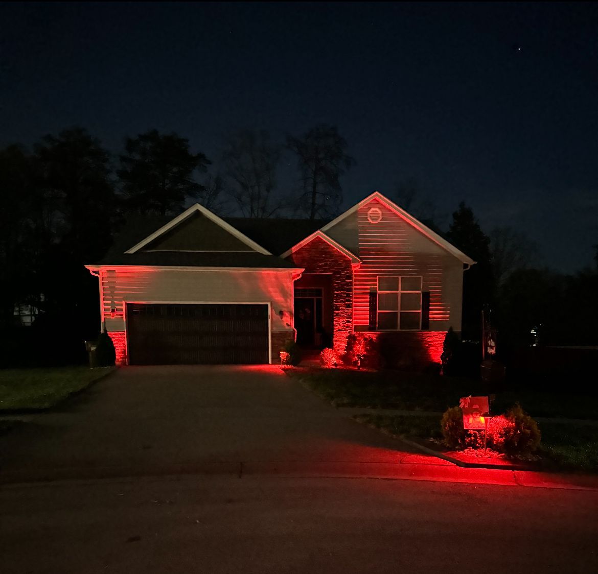 House at night lit with red lights, including the garage, porch, and landscaping.