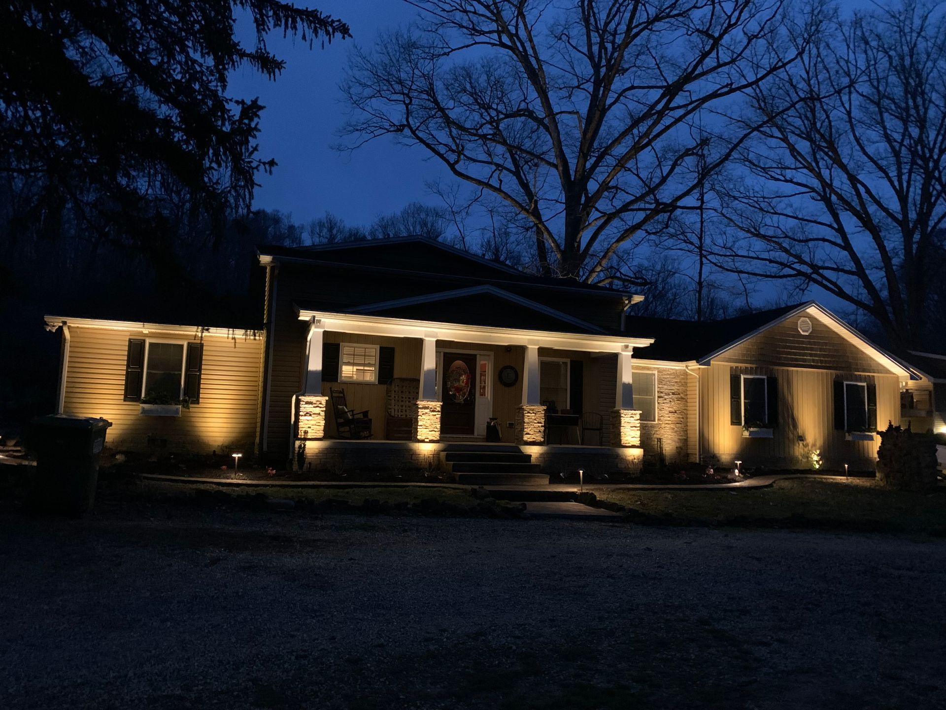 A house illuminated at night with warm, golden lighting. The porch and exterior walls are lit, set against a dark blue sky.