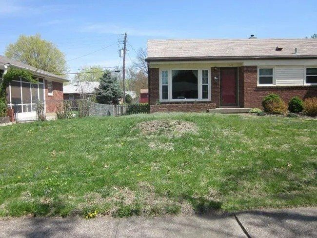 A brick and white house with a small yard on a sunny day. The yard is overgrown with grass and a small hill.
