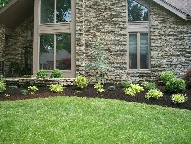 A house with stone siding and large windows, fronted by a lawn and landscaped flower beds with mulch.
