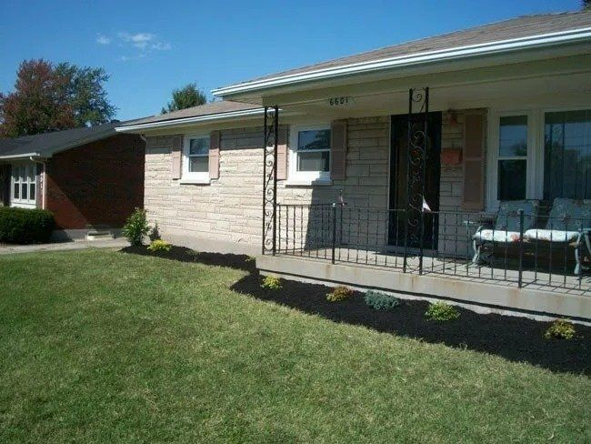 A one-story brick house with a porch and black railing. Green grass and a blue sky are in the background.