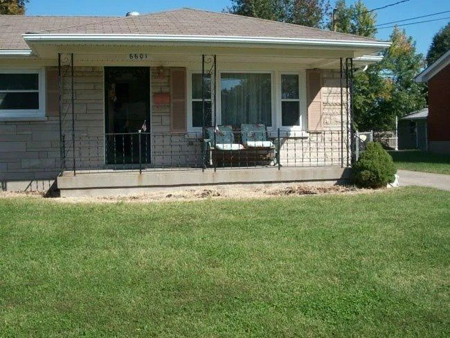 A brick bungalow with a small porch, two chairs, and a patch of grass in front of a driveway.