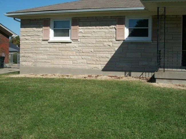 A brick house with two windows and shutters, casting a shadow on the right side, with green grass in front.