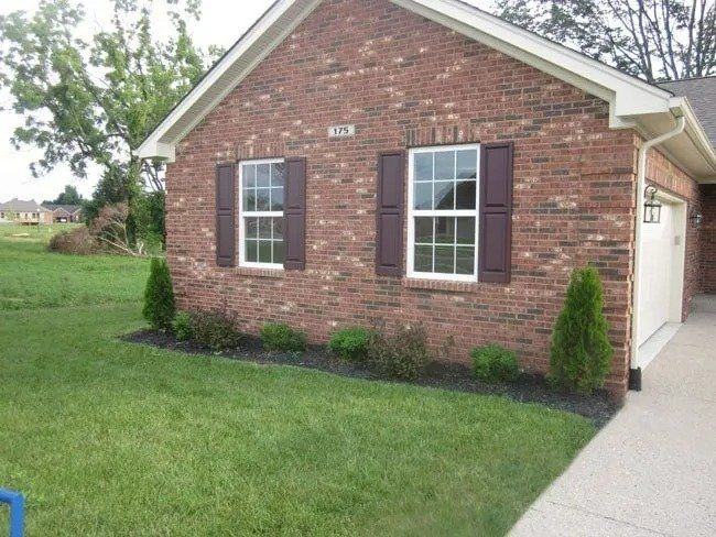 Red brick house with brown shutters and white-framed windows, small green bushes and trees in front, green lawn.