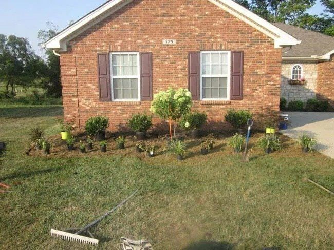 A brick house with brown shutters and a newly planted flower bed with yellow plant markers. A rake lies in the foreground.