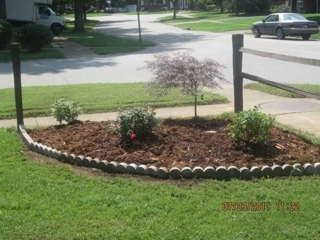 A landscaped front yard with a mulched flower bed containing small bushes and a tree, bordered by concrete blocks.