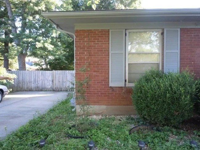 Red brick house with light blue shutters and a window. A driveway, a fence, and overgrown landscaping are in front.