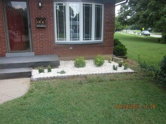 Brick house exterior with white stone flower bed and lawn. A car drives past on a road to the right.