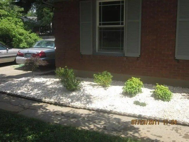 White stone bed with green bushes in front of a brick house with gray shutters. Cars are parked nearby.