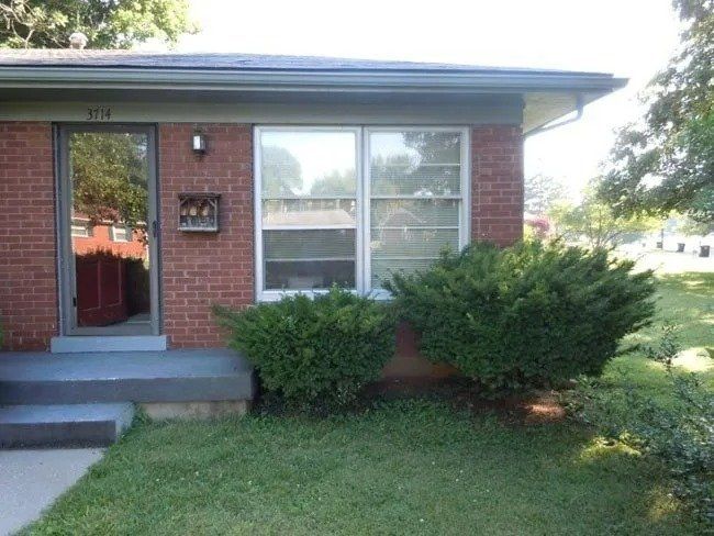 Brick house with a glass door and two windows. Two green bushes in front of the windows, with green grass in the yard.