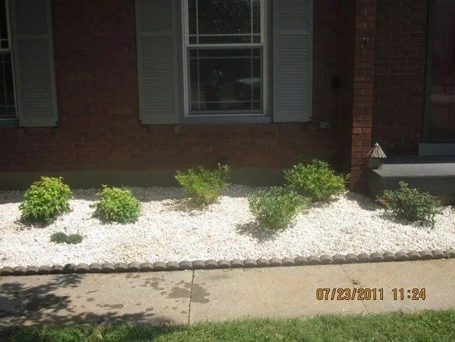 A brick home's front with white gravel landscaping, green bushes, gray shutters, and concrete path.