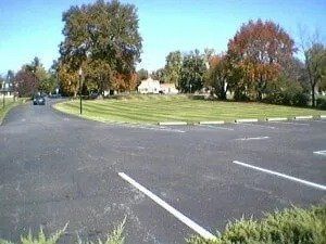 A black asphalt parking lot with white lines leads toward a grassy area with large trees, some with colorful fall foliage.