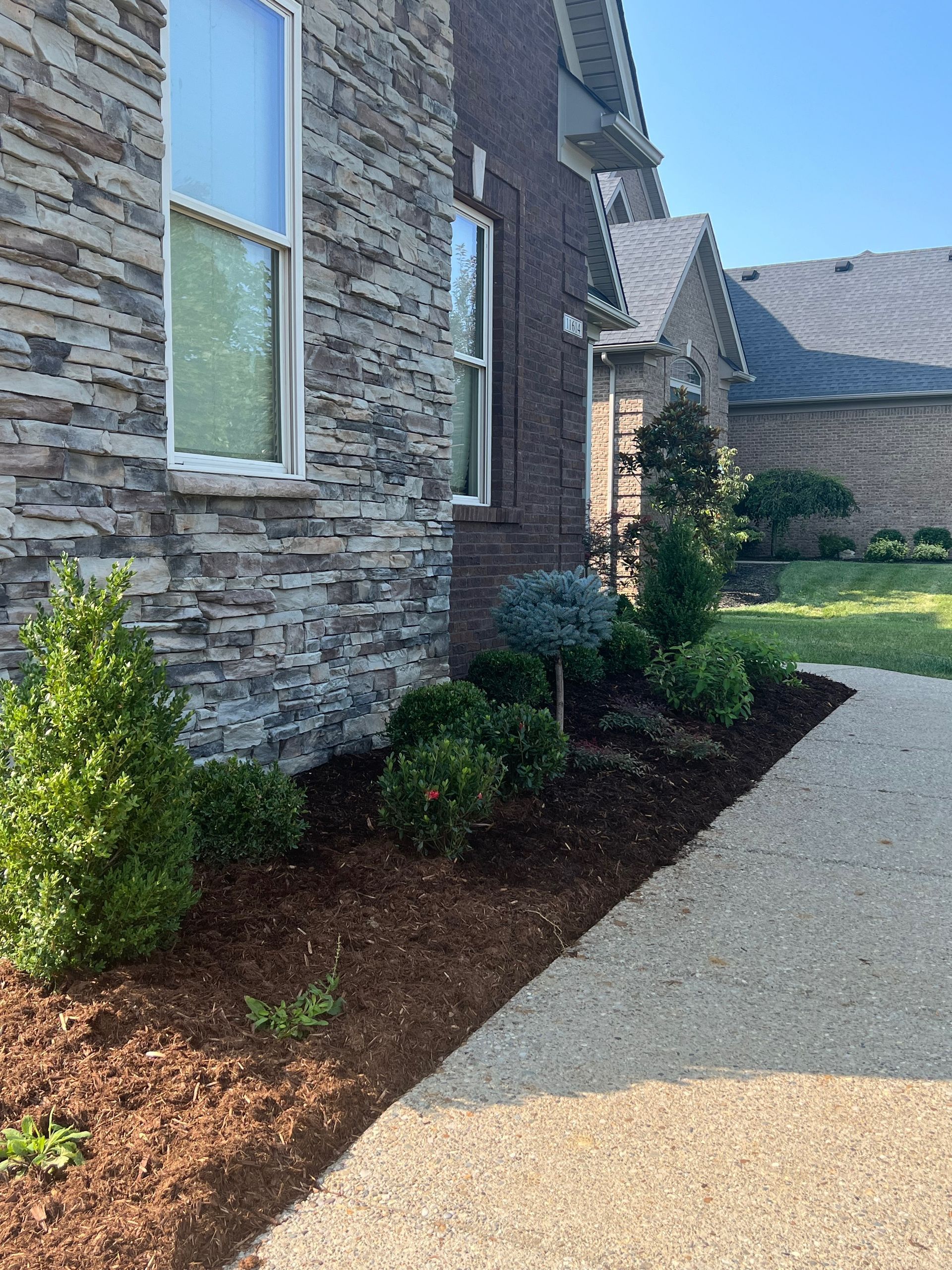 A house exterior with stone and brick walls, featuring a mulched garden bed with various green shrubs along a gravel pathway.