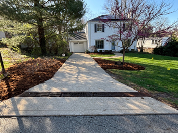 Concrete driveway leading to a white house with a garage; drainage grate in foreground, mulched borders on both sides.