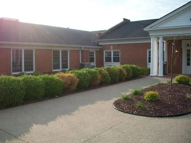 Exterior of a red brick building with a walkway lined with green and brown bushes. Sun shines on the building.