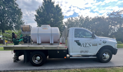 A white WCS truck with a large water tank parked on a street. A person in green work clothes stands near the tank.