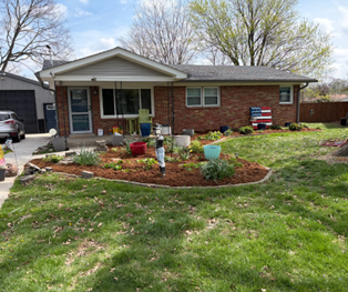 A single-story brick house with a front porch, a mulched garden bed with potted plants, and a lawn under a blue sky.
