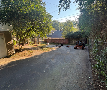 An asphalt driveway with a riding lawnmower, and debris scattered around. A brick wall and trees line the background.