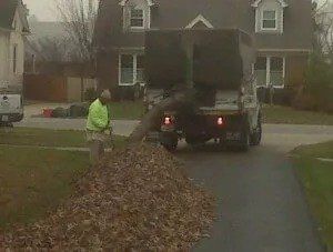 A man rakes leaves into a truck in front of a house. The truck is brown, and the man is wearing a green jacket.